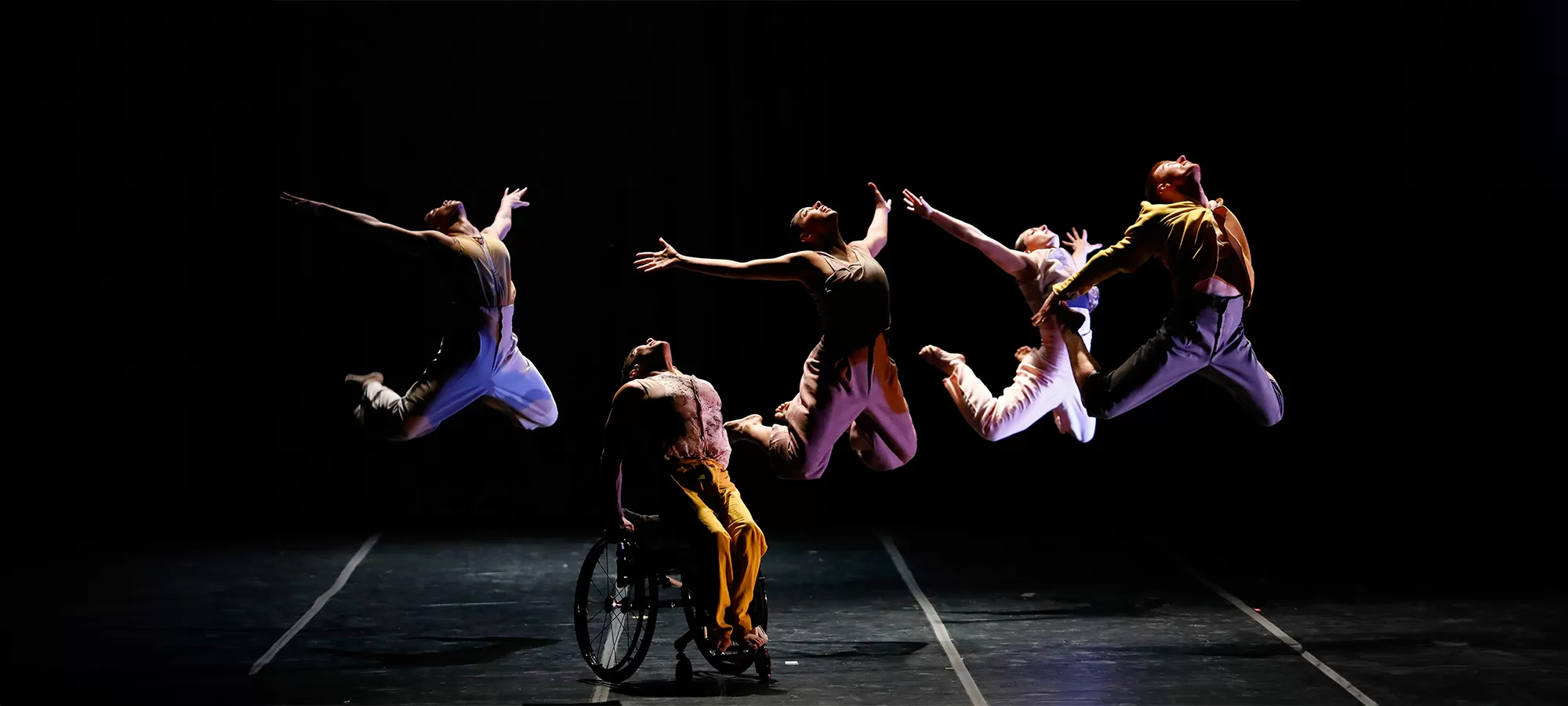 Five disabled and non-disabled dancers lift their bodies skyward, their solar plexuses rising up and out. One dancer pushes themselves out of their wheelchair with their hands, four dancers l
